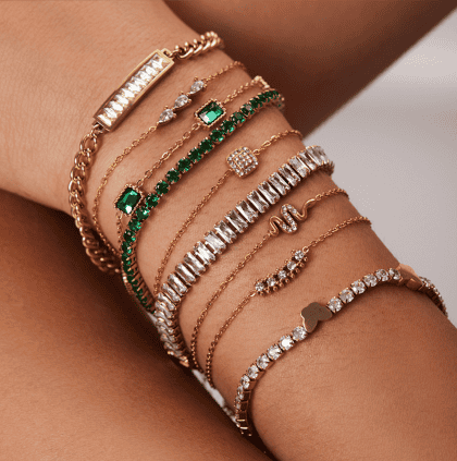 Diverse collection of custom gold and gemstone jewelry bracelets on woman's wrist, featuring emerald, diamond, and cubic zirconia designs from Nayram - Custom Jewelry.