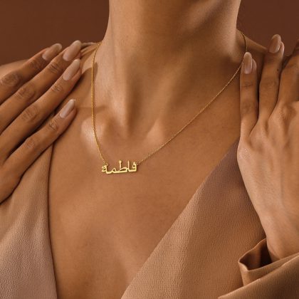 Golden personalized name necklace in Arabic script, styled with neutral-toned clothing and hands gently touching the collarbone, highlighting custom jewelry craftsmanship and elegant accessory design.